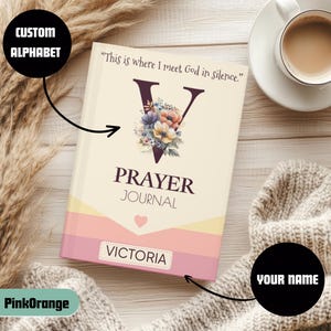 Puede incluir: Un diario de oración personalizado con el nombre "Victoria" en la portada. El diario presenta un diseño floral dentro de una gran letra "V" y el texto "Prayer Journal". La portada también incluye la cita "This is where I meet God in silence."