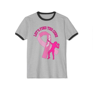 May include: Gray ringer t-shirt with black trim featuring a pink ribbon and a pink silhouette of a person throwing a ball. The text "LET'S FIND THE CURE" is arched above the ribbon.