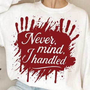 May include: White sweatshirt with a maroon graphic. The graphic features a handprint design with the words "Never mind, I handled" in a cursive font. The design has a splatter effect.