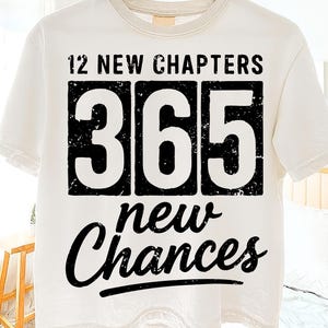 May include: White t-shirt with the text "12 NEW CHAPTERS 365 new Chances" in black. The numbers 365 are in a black rectangle. The shirt is hanging on a wooden hanger.