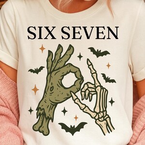 May include: Cream-colored t-shirt with the text "SIX SEVEN" in black. The shirt features a graphic of a zombie hand and a skeleton hand forming the number six and seven, with bats and stars.