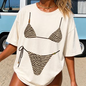 May include: An off-white short-sleeve t-shirt featuring a graphic print of a two-piece bikini with a brown and white zebra stripe pattern. The bikini top has thin straps with small brown beads, and the bikini bottom features side ties.