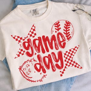 以下が含まれることがあります： クリーム色のTシャツに赤い「Game Day」のグラフィック。デザインには野球ボール、市松模様の星、野球のステッチの詳細が含まれています。シャツは青いジーンズの上に表示されています。
