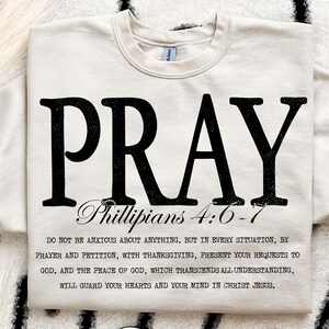 Op de afbeelding: Crèmekleurige sweatshirt met het woord "PRAY" in grote, zwarte, verweerde letters. Daaronder staat "Phillipians 4:6-7" en een langere tekst. De sweatshirt heeft een ronde hals.