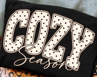 May include: A black t-shirt featuring the word COZY in large, cream-colored block letters with a dark brown polka dot pattern. Below the word COZY, the word Season is written in a cream-colored cursive script font with a thin dark brown outline.