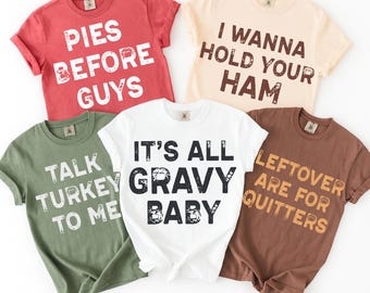 May include: Five short-sleeved t-shirts in various colors, each with a humorous Thanksgiving-themed phrase in bold white or brown text. The shirts are red, beige, white, green, and brown. Phrases include "Pies Before Guys" and "It's All Gravy Baby."