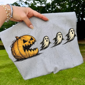 May include: A light gray t-shirt featuring a graphic print of a large, orange, jagged-toothed jack-o'-lantern chasing three small, off-white, floating ghosts. The illustration uses black ink outlines and shading to create a textured, hand-drawn appearance.