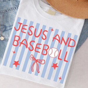 Pode incluir: Camiseta branca com as palavras "JESUS AND BASEBALL" em vermelho, com um gráfico de beisebol, estrelas vermelhas e um laço vermelho. A camisa tem listras verticais azuis.