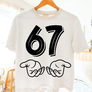 May include: White t-shirt with the number "67" in bold black font. Below the numbers are two stylized black hand illustrations. The shirt has a beige neckline and is hanging on a wooden hanger.
