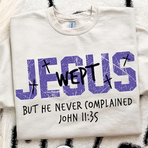 May include: Cream-colored sweatshirt with the words "JESUS WEPT" in distressed purple block letters, with small cross symbols. Below, the text reads "BUT HE NEVER COMPLAINED JOHN 11:35" in black.