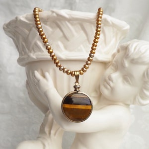 May include: A gold beaded necklace with a round pendant featuring a tiger's eye stone.