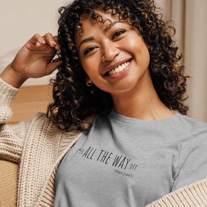 May include: A light gray long-sleeve shirt with the text "ALL THE WAY OFF" and the name "-Manjeet" in black. The shirt is worn with a beige cardigan.