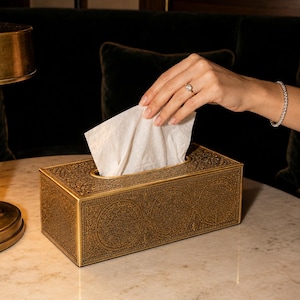 Pode incluir: Um suporte retangular para caixa de lenços com acabamento em ouro envelhecido e padrões florais e geométricos complexos em relevo em toda a superfície. A caixa contém um lenço de papel branco que sobressai pela abertura.