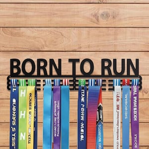 以下が含まれることがあります： 「BORN TO RUN」の黒い金属製メダルディスプレイラック。カラフルなリボンが下にぶら下がっています。ラックは木製の背景に取り付けられています。リボンにはテキストとデザインがあります。