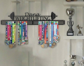 May include: Black metal weightlifting medal display with the name "Dana" and the word "WEIGHTLIFTING". The display holds numerous colorful medals with ribbons. Shelves above and below hold trophies and decorative items.