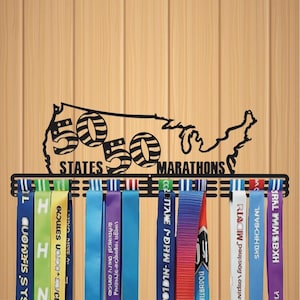 May include: Black metal marathon medal display featuring the outline of the United States with the text "50 States 50 Marathons." Colorful ribbons hang below, displaying marathon names and logos. The display is mounted on a light brown wooden surface.