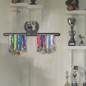 Peut inclure: Un présentoir à médailles en métal noir avec deux rangées de médailles colorées suspendues. Au-dessus du présentoir, un panneau métallique avec un logo de bouledogue et les mots "DEVIL DOGS". Le mur est blanc, et il y a des étagères avec des trophées et des figurines.