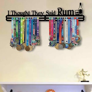 May include: Black metal medal display with the text "I Thought They Said Rum" and a bottle and glass silhouette. Colorful medals hang from the display, showcasing various race ribbons and designs. A decorative piece for runners.