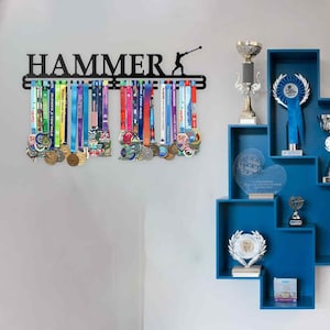 Puede incluir: Expositor de medallas de metal negro con la palabra "HAMMER" y la silueta de una persona balanceando un martillo. El expositor contiene numerosas medallas con cintas de colores. Estanterías azules a la derecha con trofeos y premios.