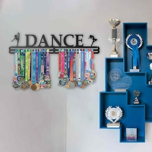 May include: Black metal medal display with the word "DANCE" and dancer silhouettes. The display holds numerous colorful medals with ribbons. Blue wall shelves hold trophies and awards, creating a motivational display.