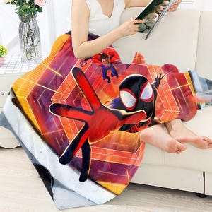 May include: A colorful blanket featuring a graphic of Spider-Man in a dynamic pose. The blanket has a geometric background with shades of orange, red, and purple. The design includes other superhero figures. The blanket is draped over a person.
