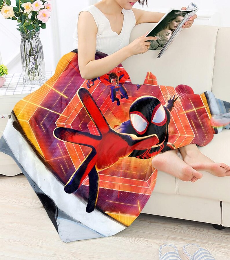 May include: A colorful blanket featuring a graphic of Spider-Man in a dynamic pose. The blanket has a geometric background with shades of orange, red, and purple. The design includes other superhero figures. The blanket is draped over a person.