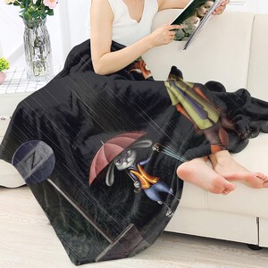 Może przedstawiać: Czarny polarowy koc z kresk&oacute;wkową ilustracją kr&oacute;lika trzymającego parasol w deszczu. Koc ma r&oacute;wnież znak Zootopii i postać w garniturze. Koc jest narzucony na osobę.