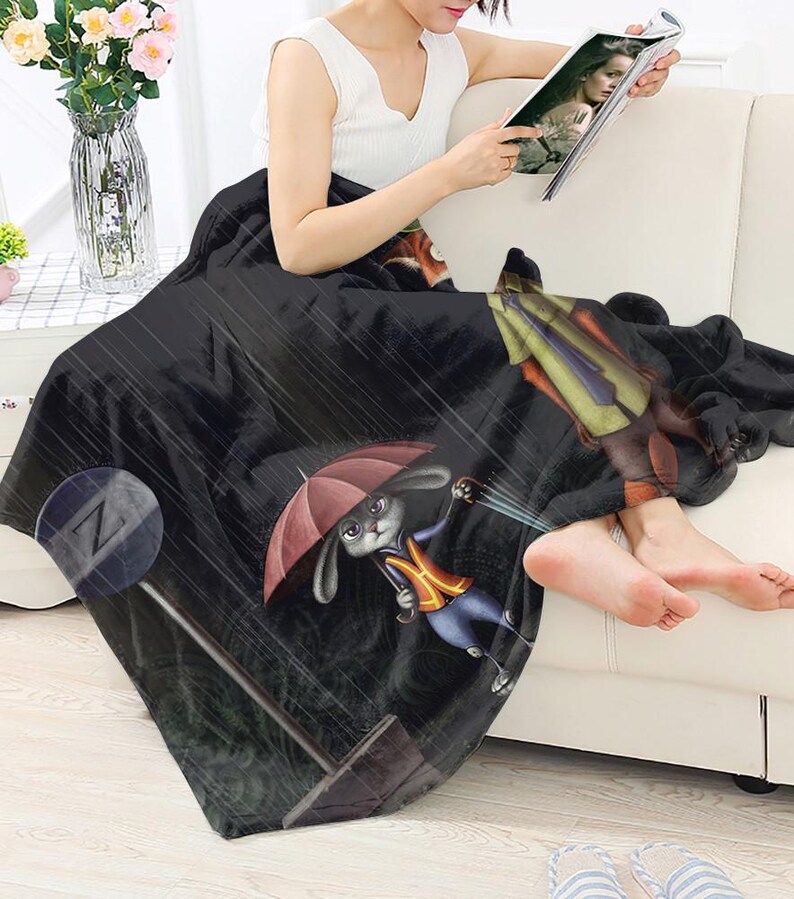 Może przedstawiać: Czarny polarowy koc z kresk&oacute;wkową ilustracją kr&oacute;lika trzymającego parasol w deszczu. Koc ma r&oacute;wnież znak Zootopii i postać w garniturze. Koc jest narzucony na osobę.