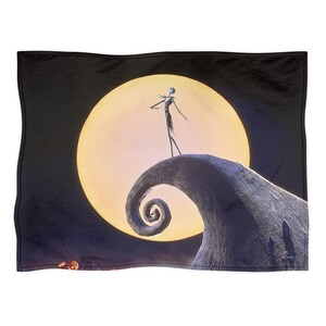 May include: Black throw blanket featuring a scene from The Nightmare Before Christmas. Jack Skellington stands on a hill in front of a large, yellow moon. The blanket has a black border and a detailed, colorful image.