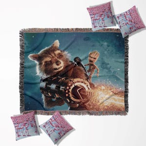 May include: A woven throw blanket and four throw pillows featuring Rocket Raccoon and Groot from Guardians of the Galaxy. The blanket has a fringed edge and a blue background. The pillows have a pink and blue design.