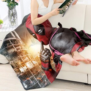 May include: A red and black Deadpool-themed blanket draped over a white couch. The blanket features a detailed graphic of Deadpool's face and costume, with a cityscape background. The blanket is soft and cozy.