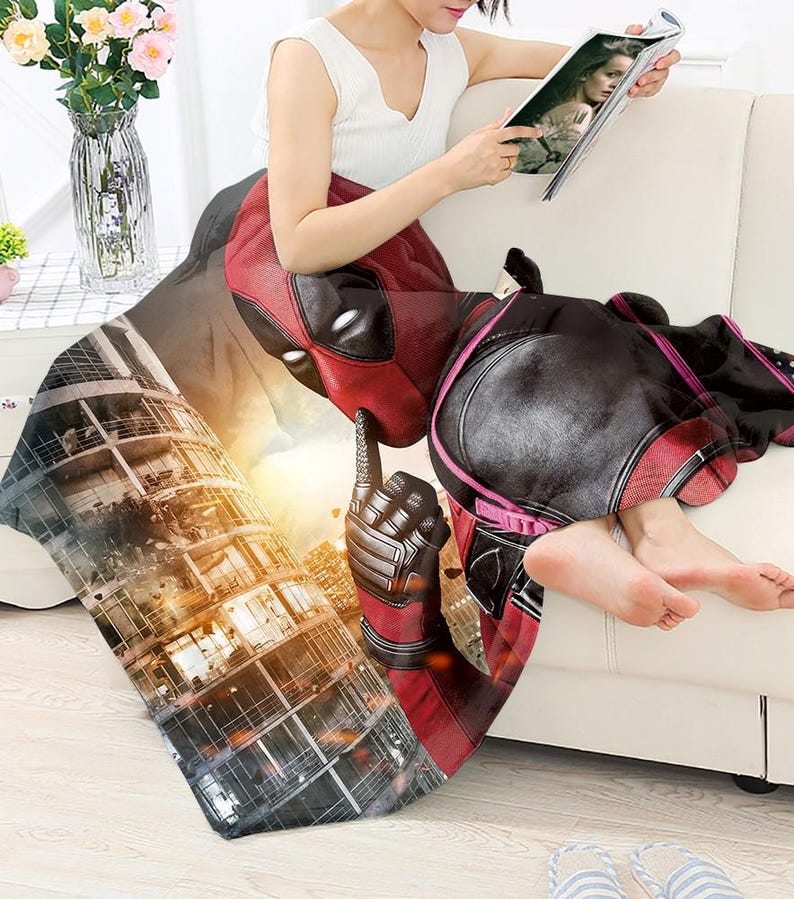 May include: A red and black Deadpool-themed blanket draped over a white couch. The blanket features a detailed graphic of Deadpool's face and costume, with a cityscape background. The blanket is soft and cozy.