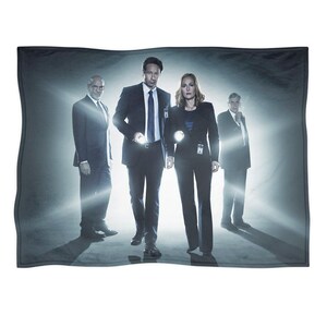 May include: A rectangular throw blanket featuring a promotional image of four people in suits, lit by bright spotlights. The image is in shades of gray and black, with the figures in the center. The blanket is made of a soft material.