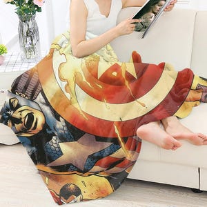 Peut inclure: Une couverture d&eacute;corative sur le th&egrave;me de Captain America, avec un design de bande dessin&eacute;e. La couverture pr&eacute;sente le bouclier du personnage en rouge, blanc et bleu, ainsi que d'autres illustrations de bandes dessin&eacute;es. La couverture est drap&eacute;e sur une personne assise sur un canap&eacute; blanc.