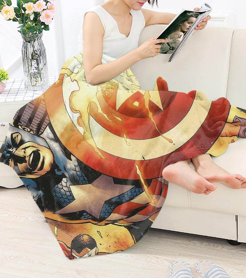 Peut inclure: Une couverture d&eacute;corative sur le th&egrave;me de Captain America, avec un design de bande dessin&eacute;e. La couverture pr&eacute;sente le bouclier du personnage en rouge, blanc et bleu, ainsi que d'autres illustrations de bandes dessin&eacute;es. La couverture est drap&eacute;e sur une personne assise sur un canap&eacute; blanc.