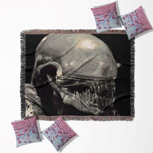 Peut inclure: Une couverture tissée noire et grise avec une image détaillée de la tête d'une créature Alien. La couverture a un bord à franges et est entourée de quatre coussins avec un motif de flamants roses roses et bleus.