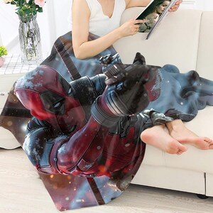 May include: A red and black Deadpool-themed blanket. The blanket features a detailed graphic of the Marvel Comics character Deadpool in a dynamic pose. The blanket is draped over a white sofa.