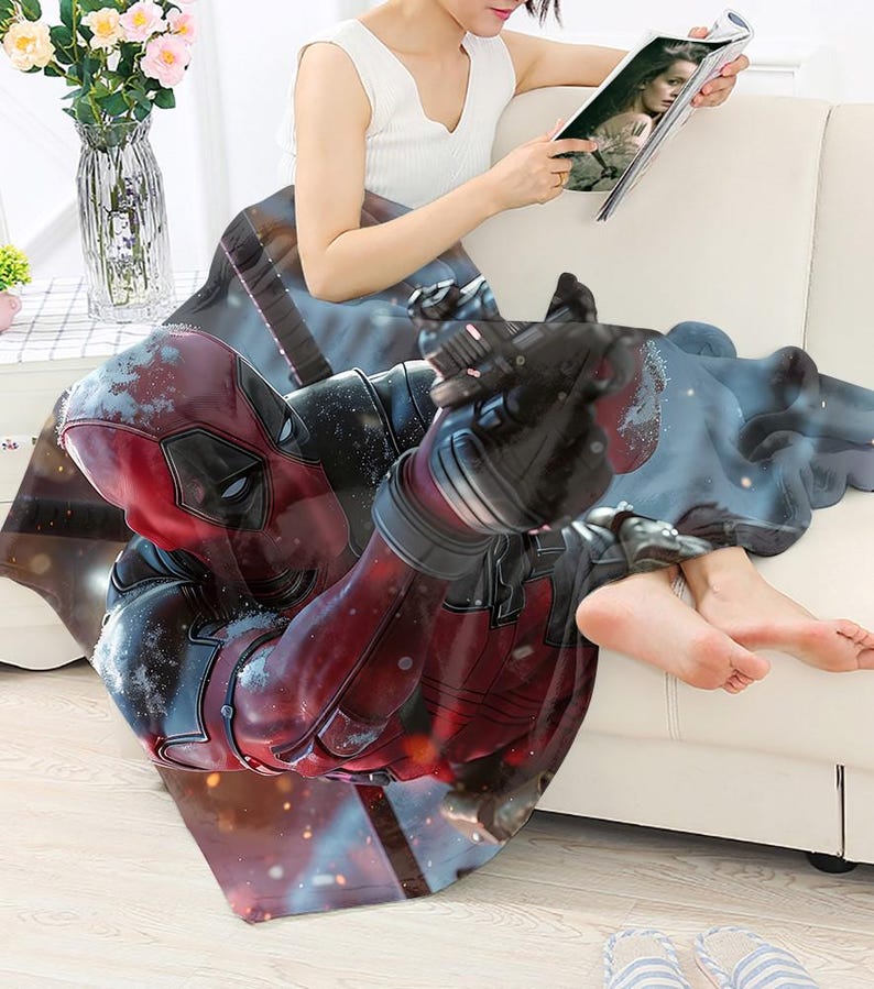 May include: A red and black Deadpool-themed blanket. The blanket features a detailed graphic of the Marvel Comics character Deadpool in a dynamic pose. The blanket is draped over a white sofa.