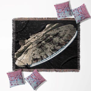 May include: A black woven throw blanket with a detailed image of a spaceship, and a black fringe. Four square throw pillows with a pink and blue flamingo design are arranged around the blanket. The spaceship is a light grey colour.