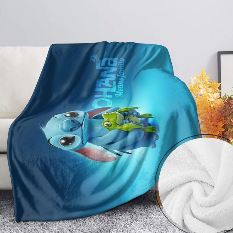 Pode incluir: Um cobertor de pel&uacute;cia azul com o personagem Stitch de Lilo & Stitch, segurando um sapo verde. O cobertor tem as palavras "OHANA Means family" impressas. O cobertor &eacute; macio e fofo.