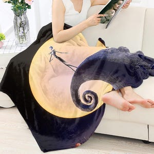 May include: A black and yellow throw blanket featuring a full moon and a silhouette of Jack Skellington from The Nightmare Before Christmas. The blanket is draped over a white sofa.