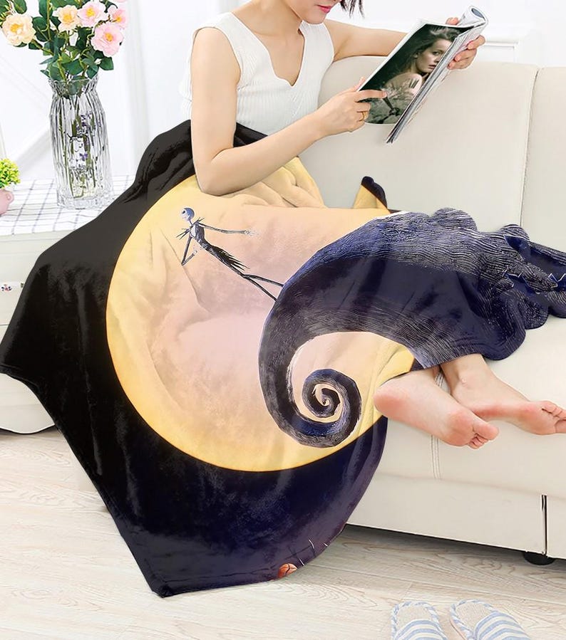 May include: A black and yellow throw blanket featuring a full moon and a silhouette of Jack Skellington from The Nightmare Before Christmas. The blanket is draped over a white sofa.