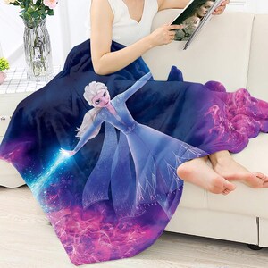 Puede incluir: Una manta de forro polar azul marino con una colorida ilustraci&oacute;n de Elsa de la pel&iacute;cula Frozen. El dise&ntilde;o incorpora tonos azules, morados y rosas. La manta est&aacute; sobre una persona sentada en un sof&aacute; blanco.
