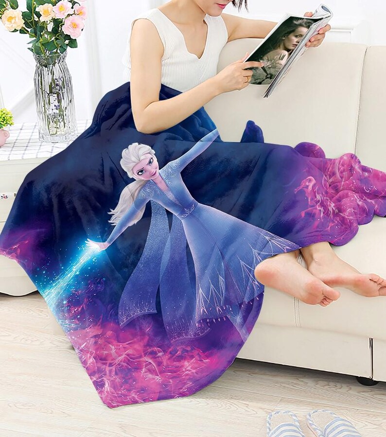 Puede incluir: Una manta de forro polar azul marino con una colorida ilustraci&oacute;n de Elsa de la pel&iacute;cula Frozen. El dise&ntilde;o incorpora tonos azules, morados y rosas. La manta est&aacute; sobre una persona sentada en un sof&aacute; blanco.