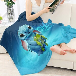 Pode incluir: Um cobertor de l&atilde; azul com uma imagem de desenho animado de Stitch segurando um sapo verde. O cobertor tem o texto "OHANA Means Family" em branco. O cobertor est&aacute; sobre uma pessoa sentada em um sof&aacute; branco.