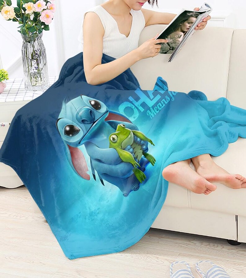 Pode incluir: Um cobertor de l&atilde; azul com uma imagem de desenho animado de Stitch segurando um sapo verde. O cobertor tem o texto "OHANA Means Family" em branco. O cobertor est&aacute; sobre uma pessoa sentada em um sof&aacute; branco.