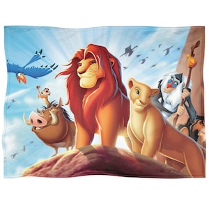 Op de afbeelding: Een rechthoekige deken met een illustratie van personages uit The Lion King. Het ontwerp toont Simba, Nala, Timon, Pumbaa, Zazu en Rafiki. De achtergrond is een blauwe lucht met vogels.