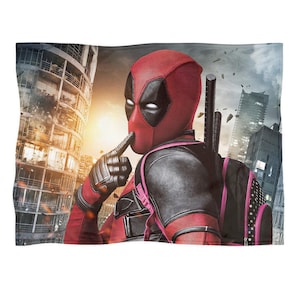 May include: A red and black Deadpool blanket featuring the character in his signature mask and costume, with a cityscape background. The blanket showcases the superhero in a dynamic pose, with a finger to his lips.