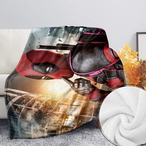 May include: A plush blanket featuring a vibrant image of the character Deadpool in his signature red and black costume. The design includes a cityscape backdrop with a soft, white, textured underside.