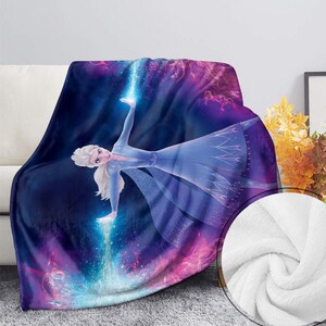 Puede incluir: Una manta suave en tonos azules y morados, que muestra un personaje de una pel&iacute;cula de animaci&oacute;n. El personaje tiene cabello rubio y un vestido azul, con un chorro de luz azul que emana de su mano. La manta est&aacute; sobre un sof&aacute; blanco.